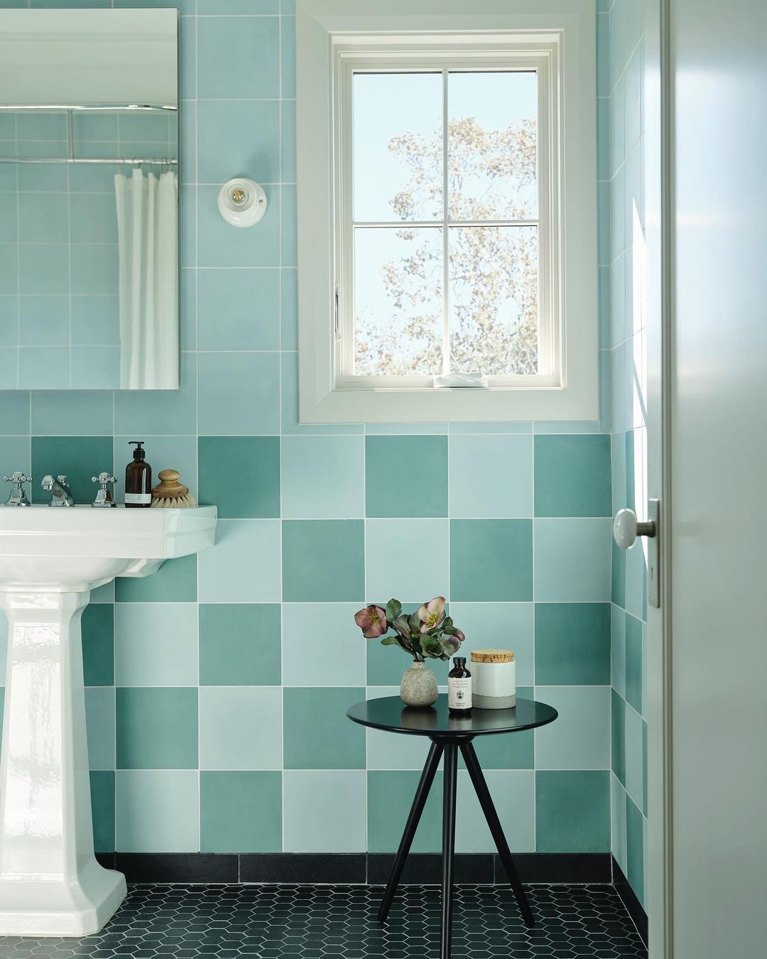 A bathroom with light blue and teal checkered tiles, white pedestal sink, hexagonal dark floor tiles, a window with a view of trees, and a round black side table holding flowers and small toiletries.