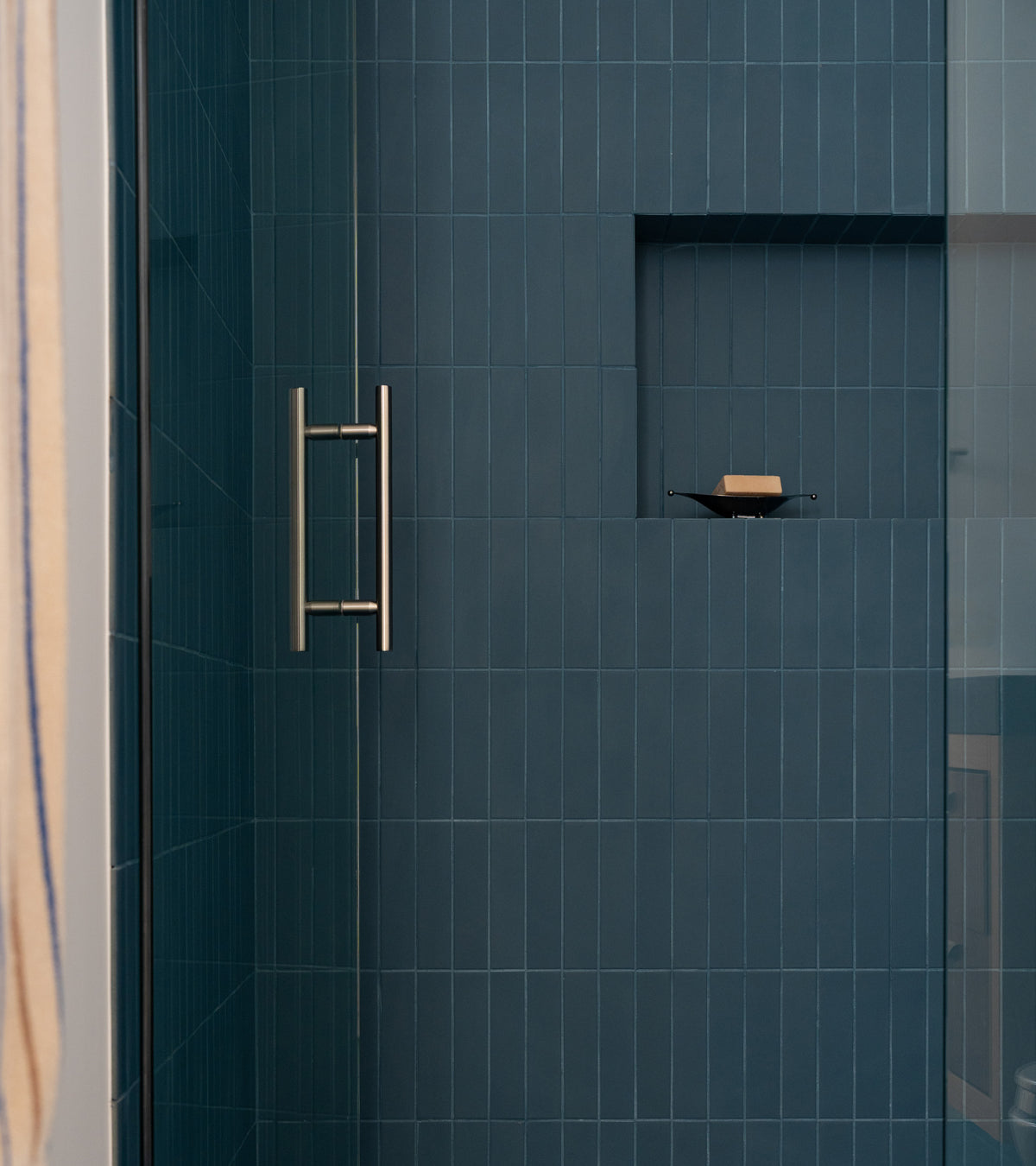 A modern shower features Zia Tile's Iconic Blue 2x8 ceramic tiles in a sleek vertical layout, a metal handle on the glass door, and a recessed shelf with a single bar of soap.