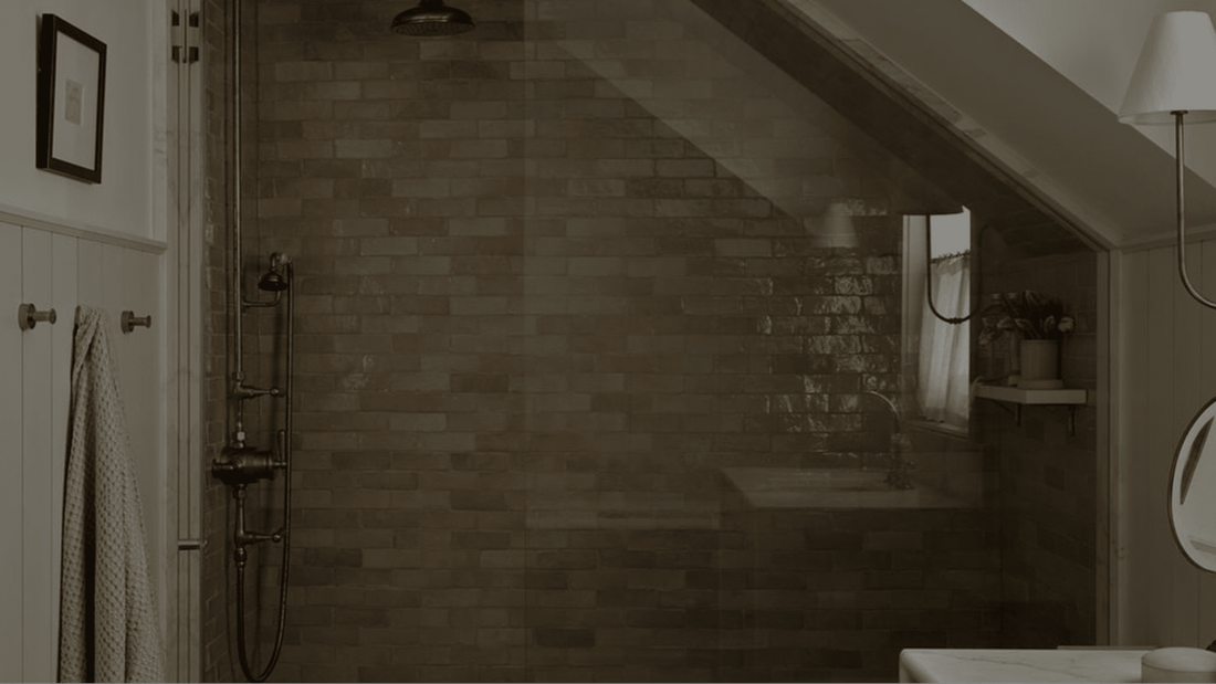 A tiled attic shower with a glass door, a rainfall showerhead, a hanging towel, a round mirror, a wall lamp, and a small window letting in light.