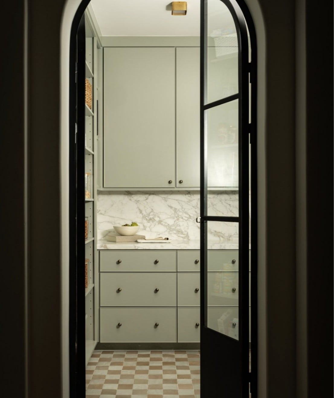 A view through an arched doorway into a modern, light green pantry with cabinets, marble backsplash, open shelving, and checkered tile floor. A bowl and kitchen items sit on the counter.