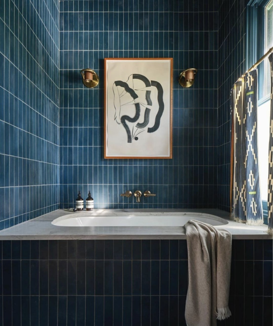 A modern bathroom with dark blue vertical tiles, a white bathtub, and a gray towel draped over the edge. Above the tub hangs abstract art, flanked by two brass wall lights. Light enters from a window with patterned curtains.