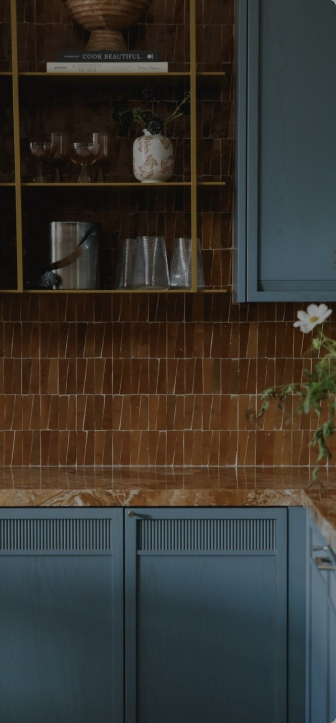 A kitchen with blue cabinets, a brown patterned tile backsplash, gold shelves holding glasses, a vase, and a book, and a marble countertop with a plant in the corner.