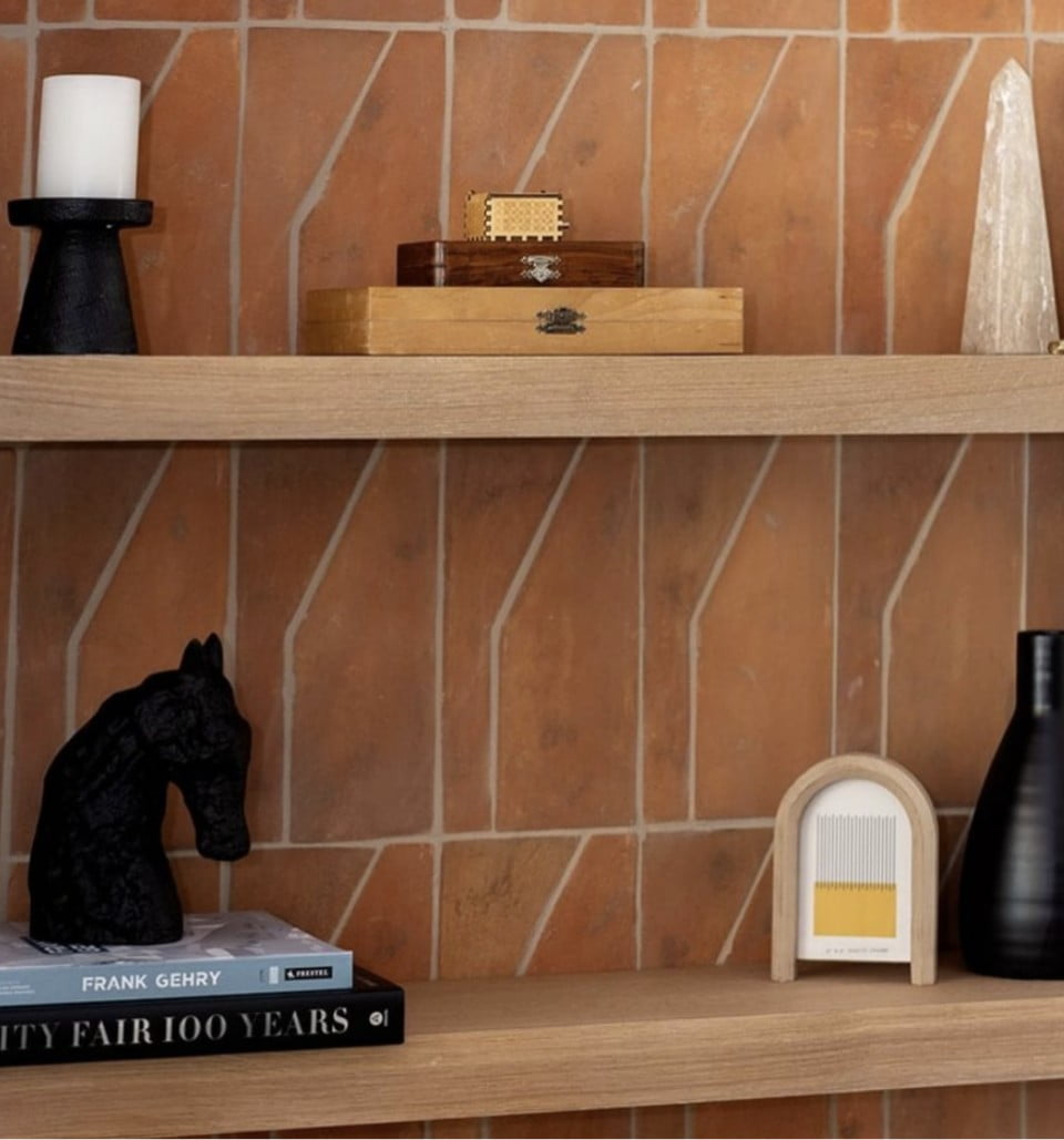 Two wooden shelves on a brick wall display a black candle holder, a wooden box, a clear crystal, a black horse head sculpture, books, a modern match holder, and a black vase.
