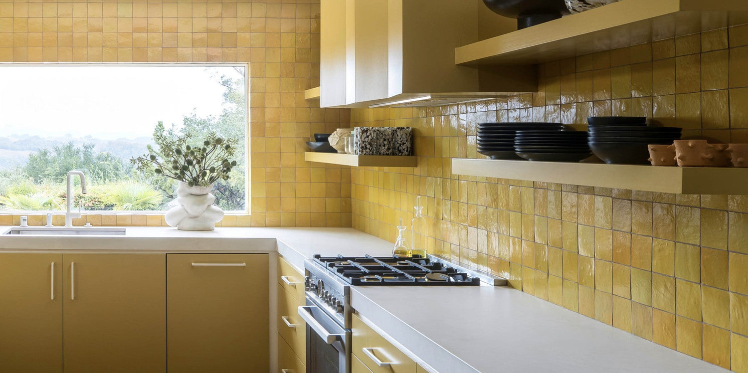 A modern kitchen with yellow square tile walls, open shelves holding dishes, a gas stove, a sink under a large window with an outdoor view, and a light-colored countertop.