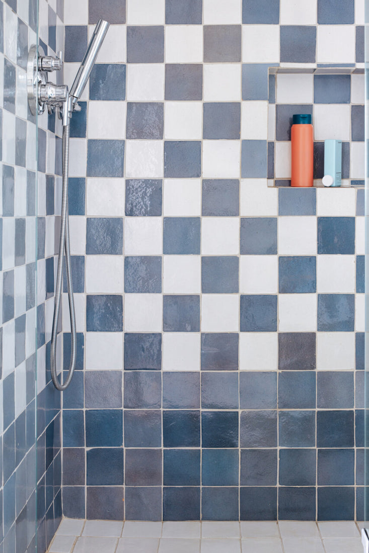 A shower with blue and white checkered tiles, a silver handheld showerhead, and a recessed shelf holding blue and orange bottles.