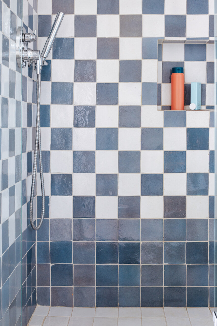 A shower with blue and white checkered tiles, a silver handheld showerhead, and a recessed shelf holding blue and orange bottles.