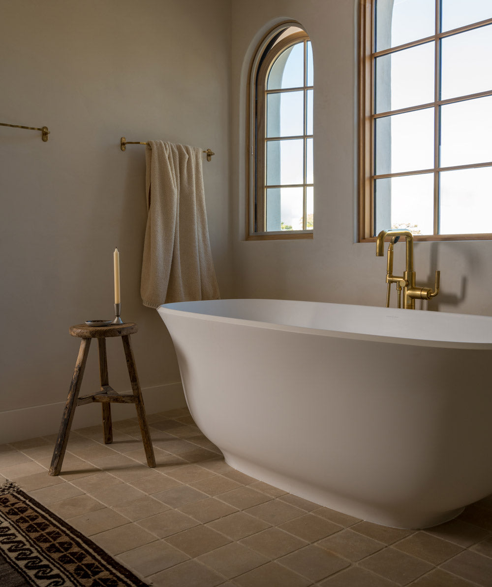 A minimalist bathroom with a white freestanding tub, arched windows, a towel hanging on a brass rod, and a rustic wooden stool holding a candle. The room has beige tiles and soft natural light.