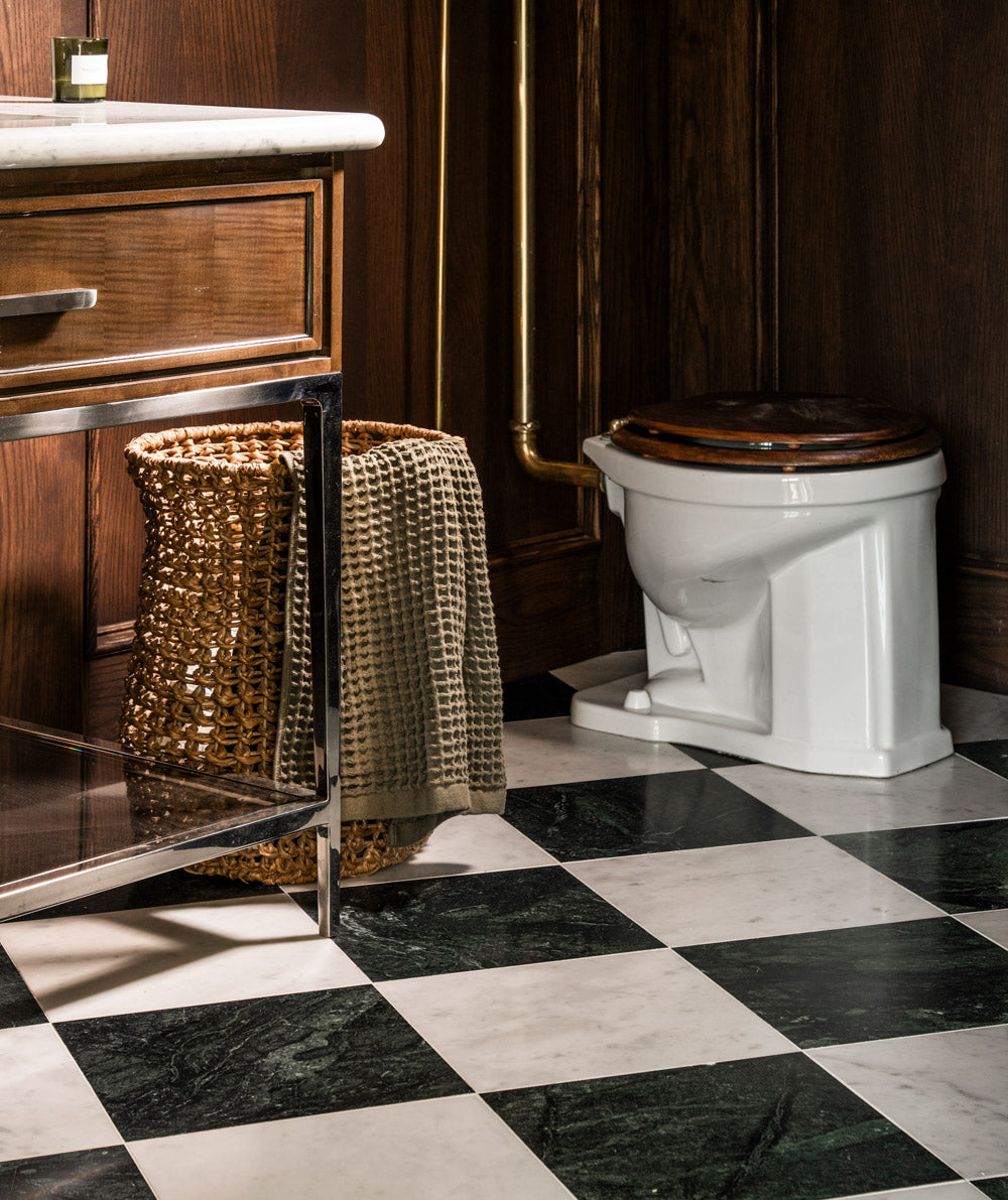 A bathroom corner with a checkered black and white tile floor, a wooden vanity with a marble top, a woven laundry basket, and a white toilet with a dark wooden seat.