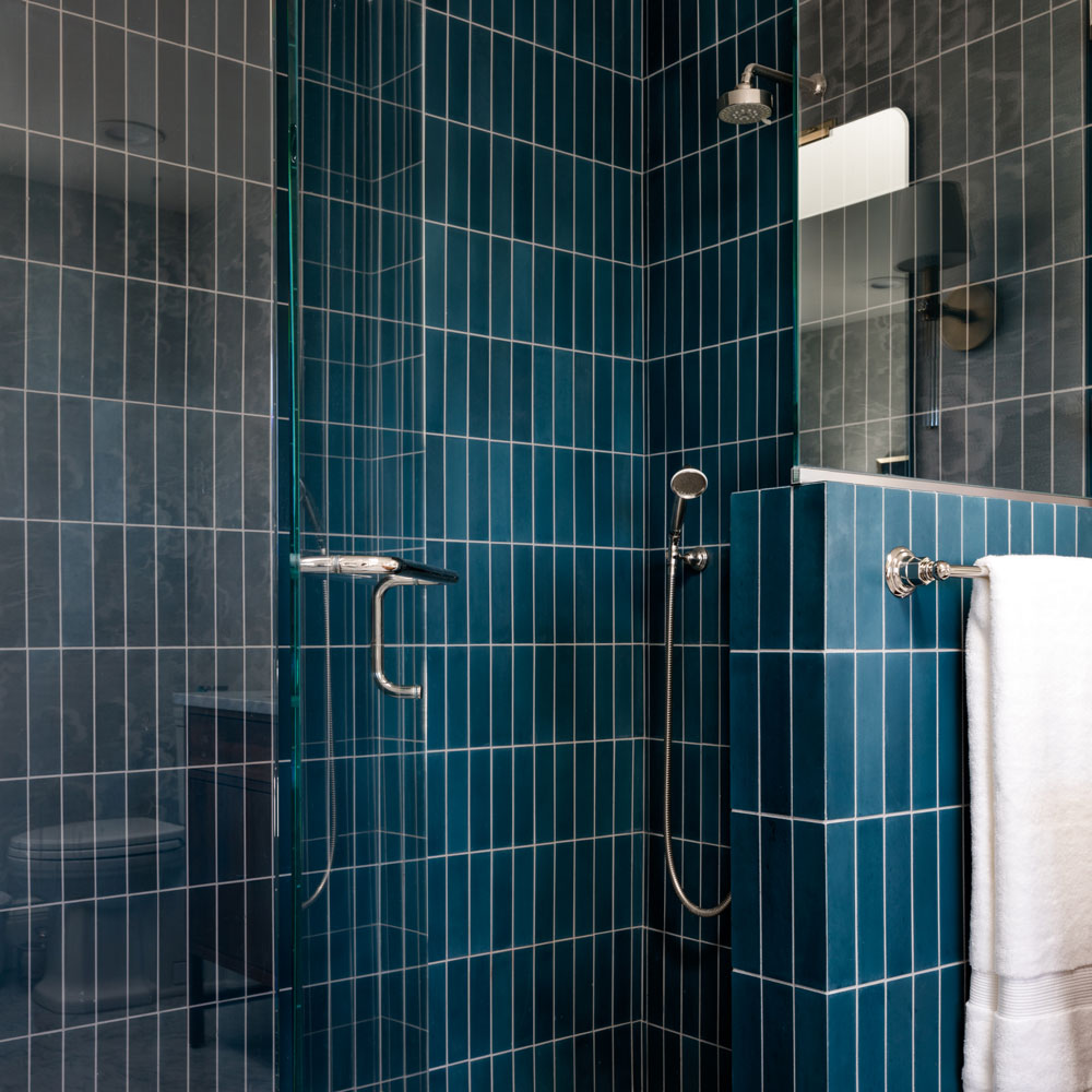A modern shower with blue vertical rectangular tiles, a glass door, a silver showerhead, and a handheld shower. A white towel hangs on a rack on a short partition wall next to the shower.