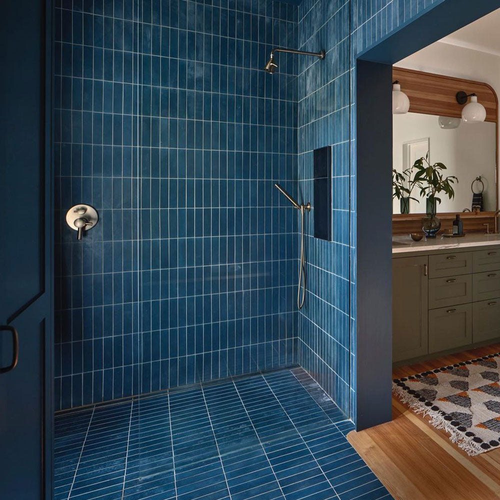 Modern bathroom with a walk-in shower featuring vertical blue tiles, a silver showerhead, and a wooden vanity with a mirror and plants visible outside the shower area.