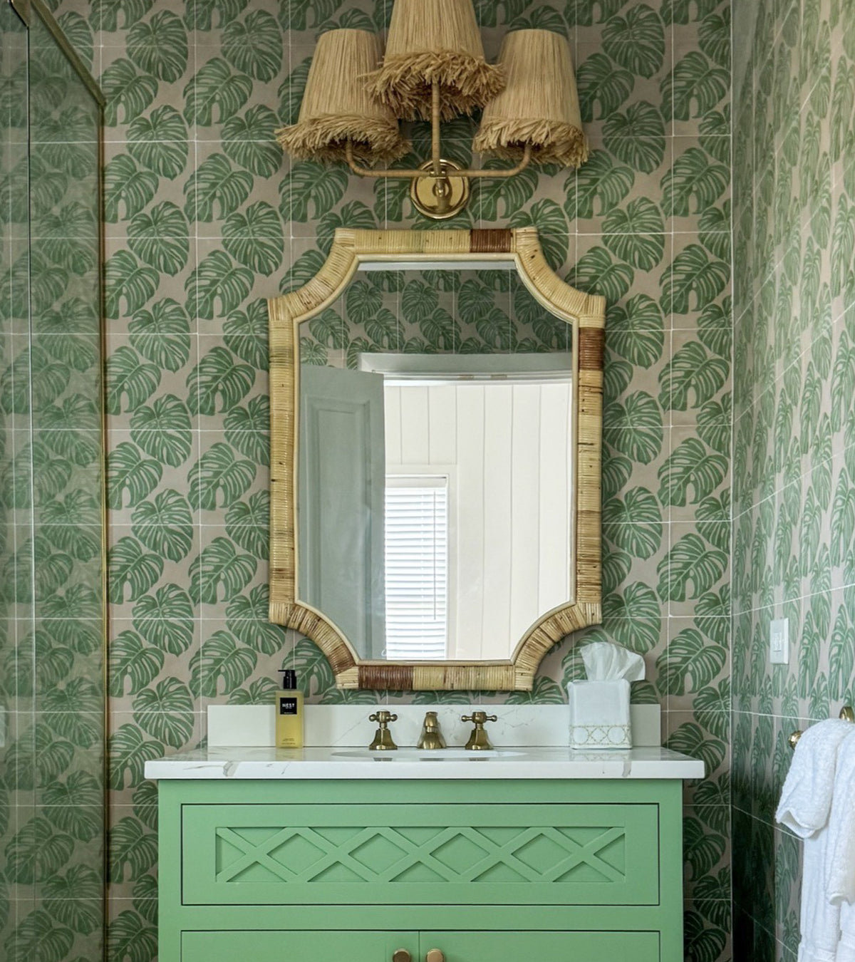 A bathroom features green leaf-patterned wallpaper inspired by Monstera 8x8 tiles by vendor-unknown, a gold-framed mirror above a mint green vanity with white countertop, brass faucets, tissue box, and three-shade wicker wall sconces.