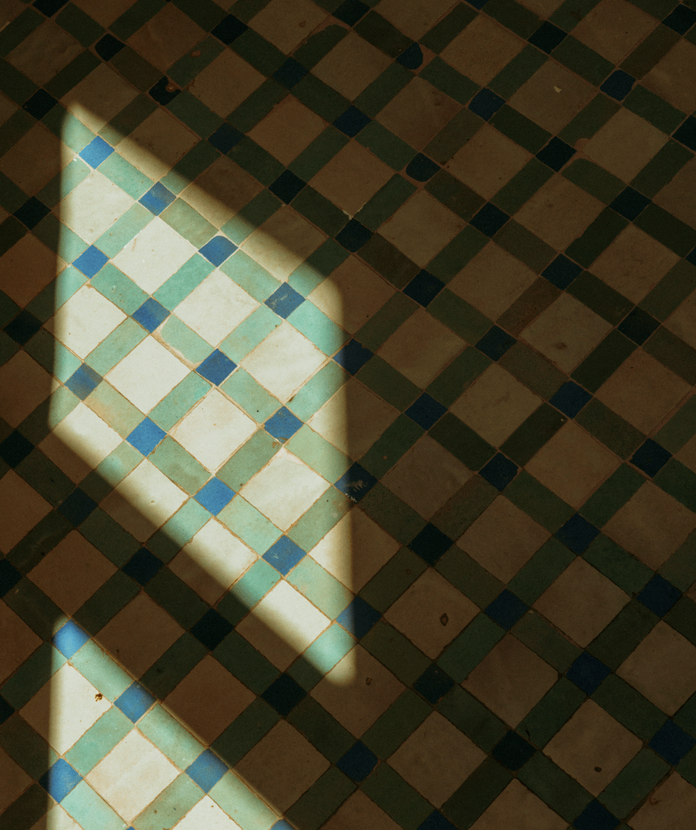 Sunlight casts a rectangular patch of light onto a tiled floor with a blue and green diamond pattern, creating contrast between the bright area and the surrounding shadows.