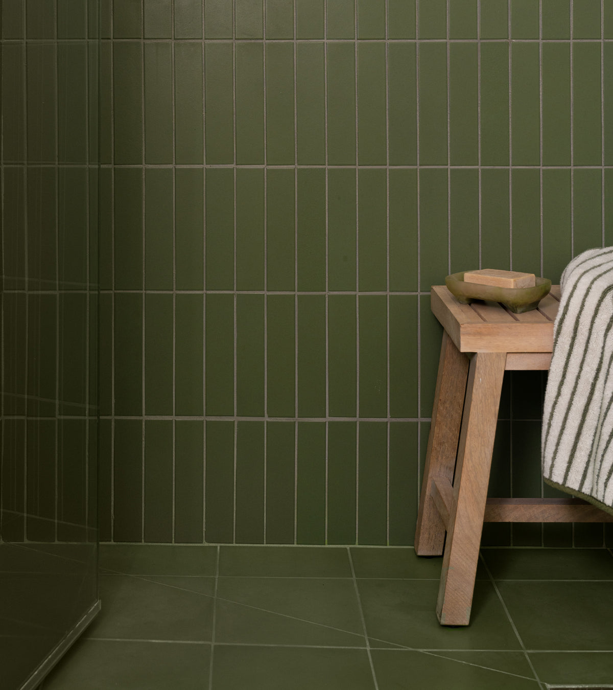 A bathroom features olive green rectangular wall tiles and Olivine 8x8 cement tile flooring by vendor-unknown. In the corner, a light wooden stool holds a striped towel and a small dish.