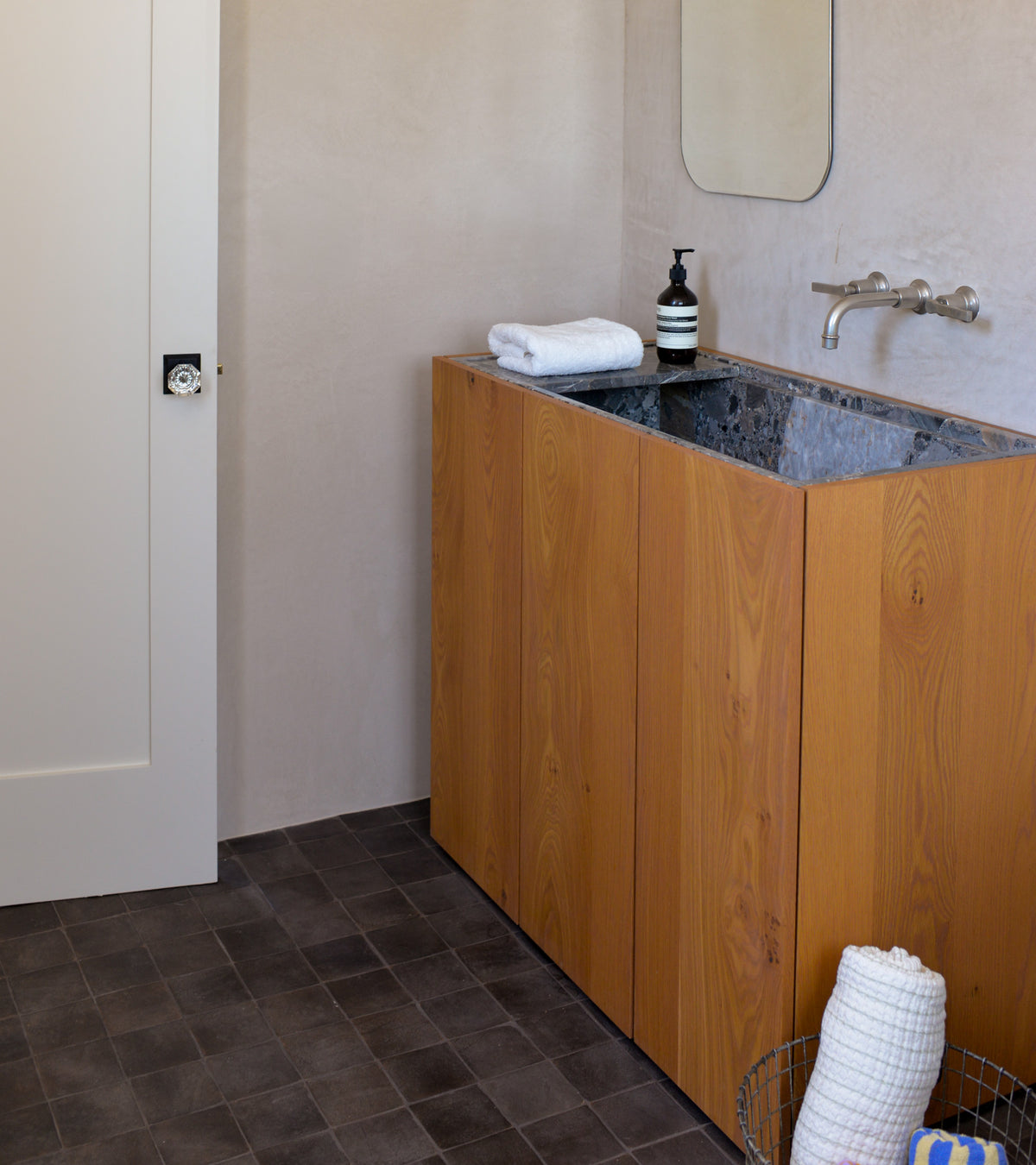 A modern bathroom features a wooden vanity, dark stone countertop, integrated sink, and wall-mounted faucet. Rolled white towels sit on the counter and in a wire basket atop Zia Tile's 4x4 Square + Oscura handmade tiles.