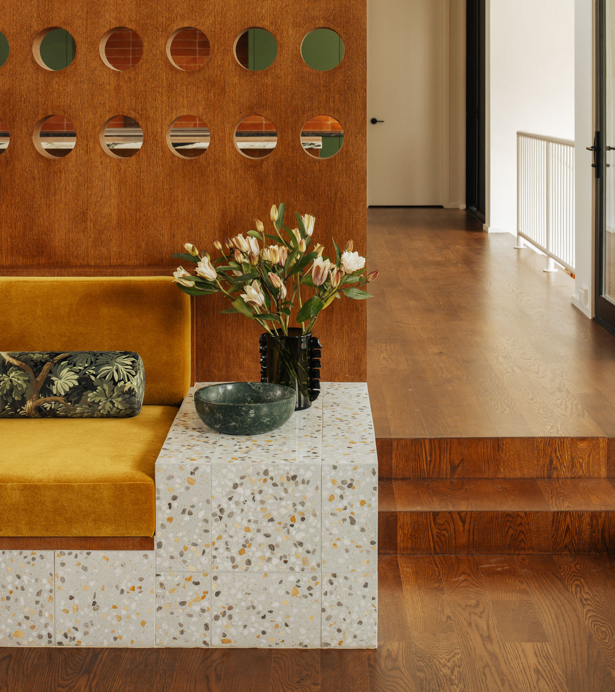 A modern interior features a mustard yellow bench with a floral cushion, a Zia Tile Parker 12x12 terrazzo cement tile side table topped with a green bowl and flowers, plus a wooden partition with round cutouts and brown wood floors.