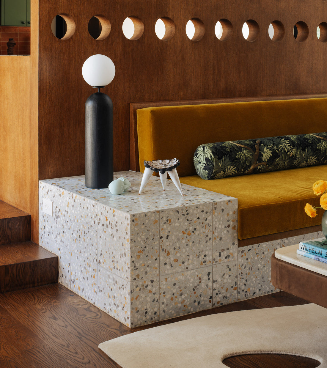 A modern living room with a terrazzo side table, black and white lamp, small bowl, mustard-yellow velvet sofa, wooden partition with circular cutouts, floral cushion, and cement flooring by Zia Tile's Parker 12x12 completes the stylish setting.