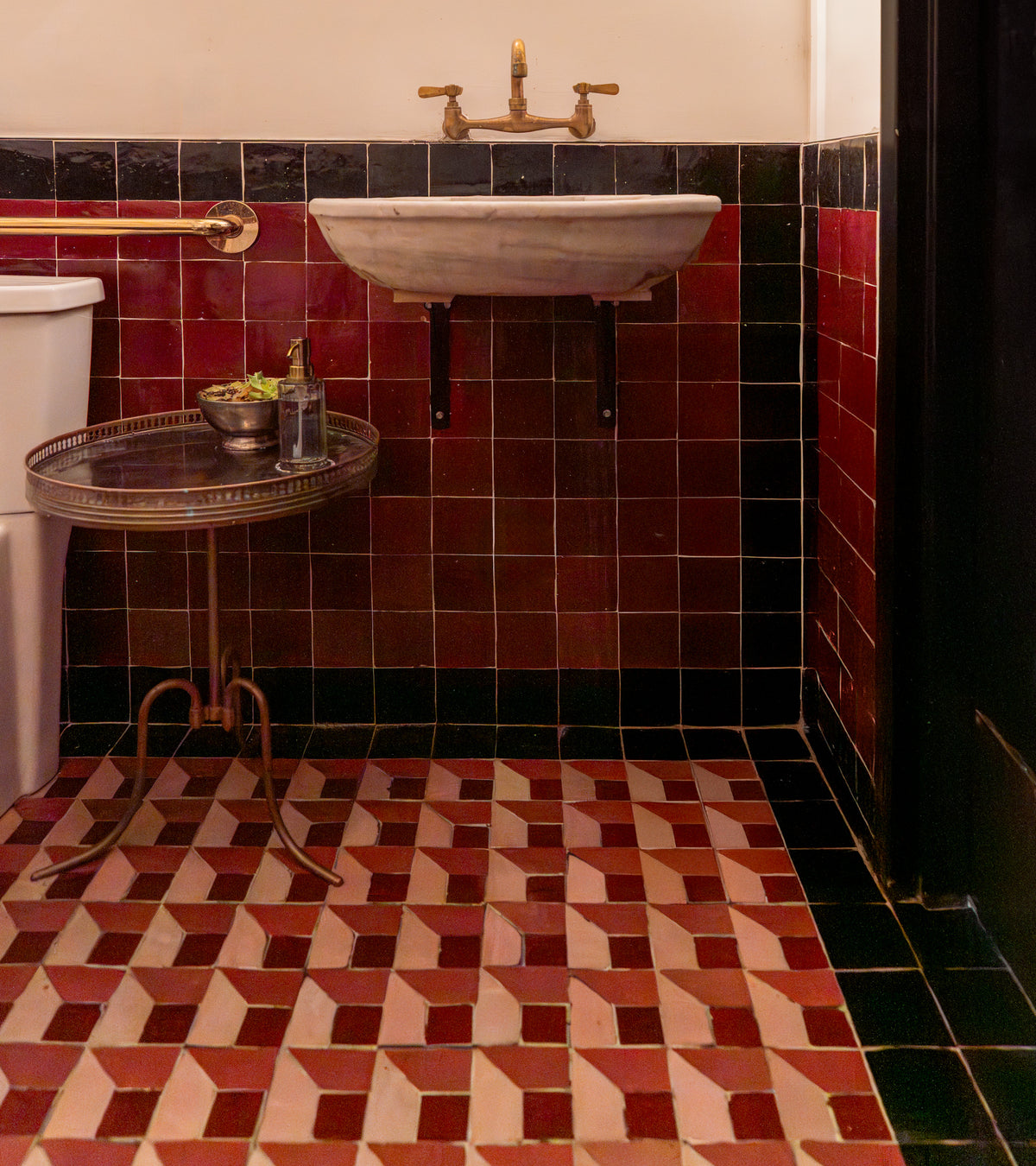 A vintage bathroom with a pedestal sink, gold faucet, a small round table with soap and a plant, black and burgundy wall tiles, and cream, red, and black Perpetual Check 3 patterned floor tiles by Zia Tile reminiscent of Moroccan design.