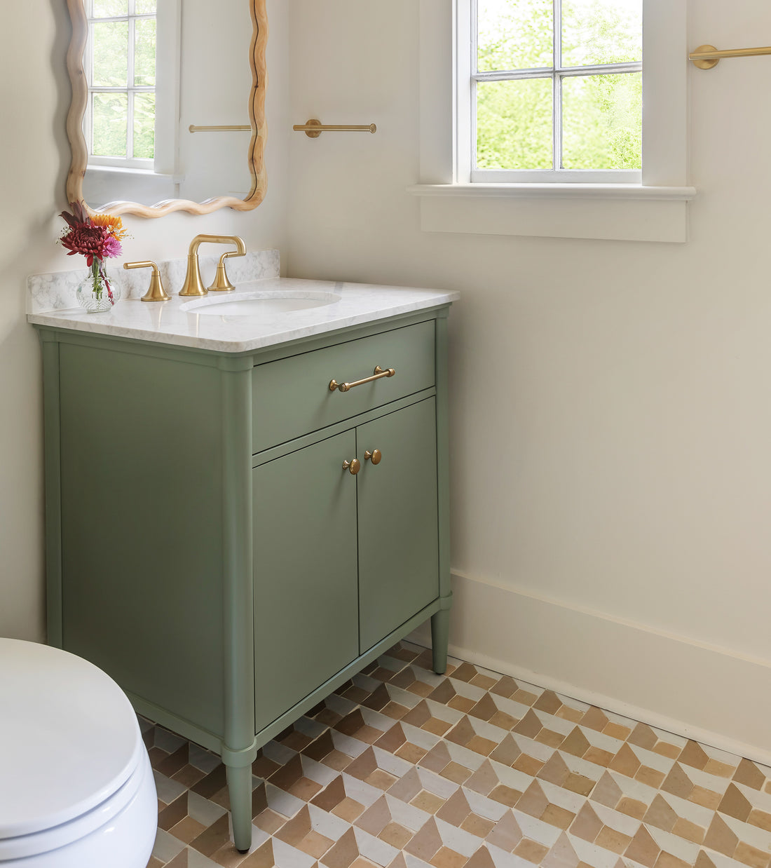 A small bathroom features a green vanity with gold hardware, marble countertop, gold faucet, decorative mirror with gold frame, a single red flower in a vase, and Zia Tile’s Radian Inline 1 geometric patterned Moroccan tile floor.