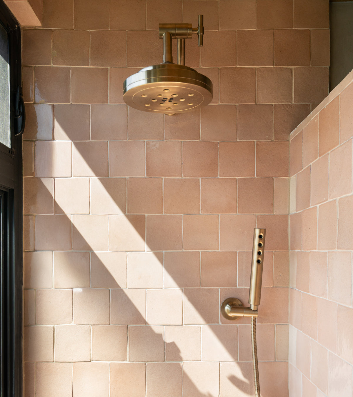 A modern shower with a brass showerhead and handheld sprayer is mounted on a wall of Zia Tile's Posada 4x4 Square tiles. Sunlight highlights the handmade cotto, adding San Miguel de Allende charm to the space.