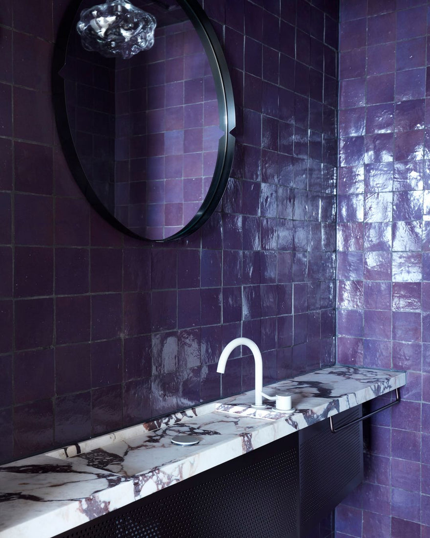 A modern bathroom with glossy dark purple tiled walls, a round mirror, a white marble countertop with dark veining, a sleek white faucet, and a minimalist soap dish.