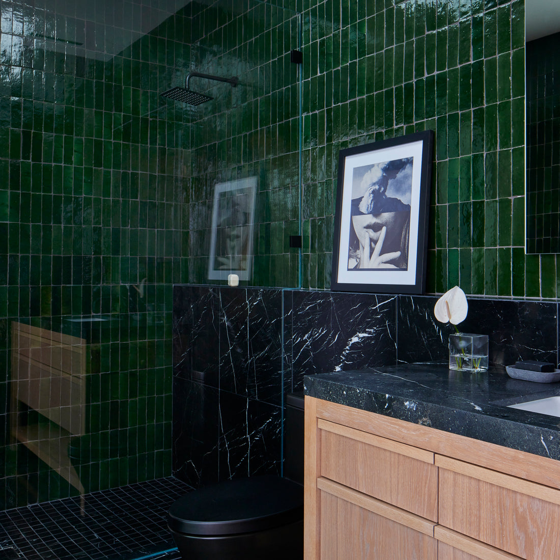 A modern bathroom with glossy green tile shower walls, black marble floors and backsplash, a black toilet, light wood vanity with a black countertop, and a framed photo resting on the marble ledge.
