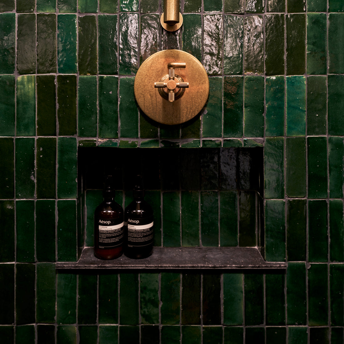 A shower with glossy dark green square tiles, a brass shower control fixture, and a built-in shelf holding two bottles of Aesop products.