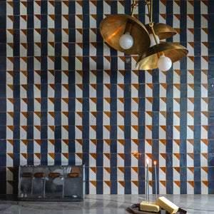A Radian Inline 2 geometric kitchen backsplash by Zia Tile sets the scene, paired with a gold three-bulb pendant, marble countertop, two lit candles, a tray with gold bars, and a vintage metal box with leather straps in the foreground.