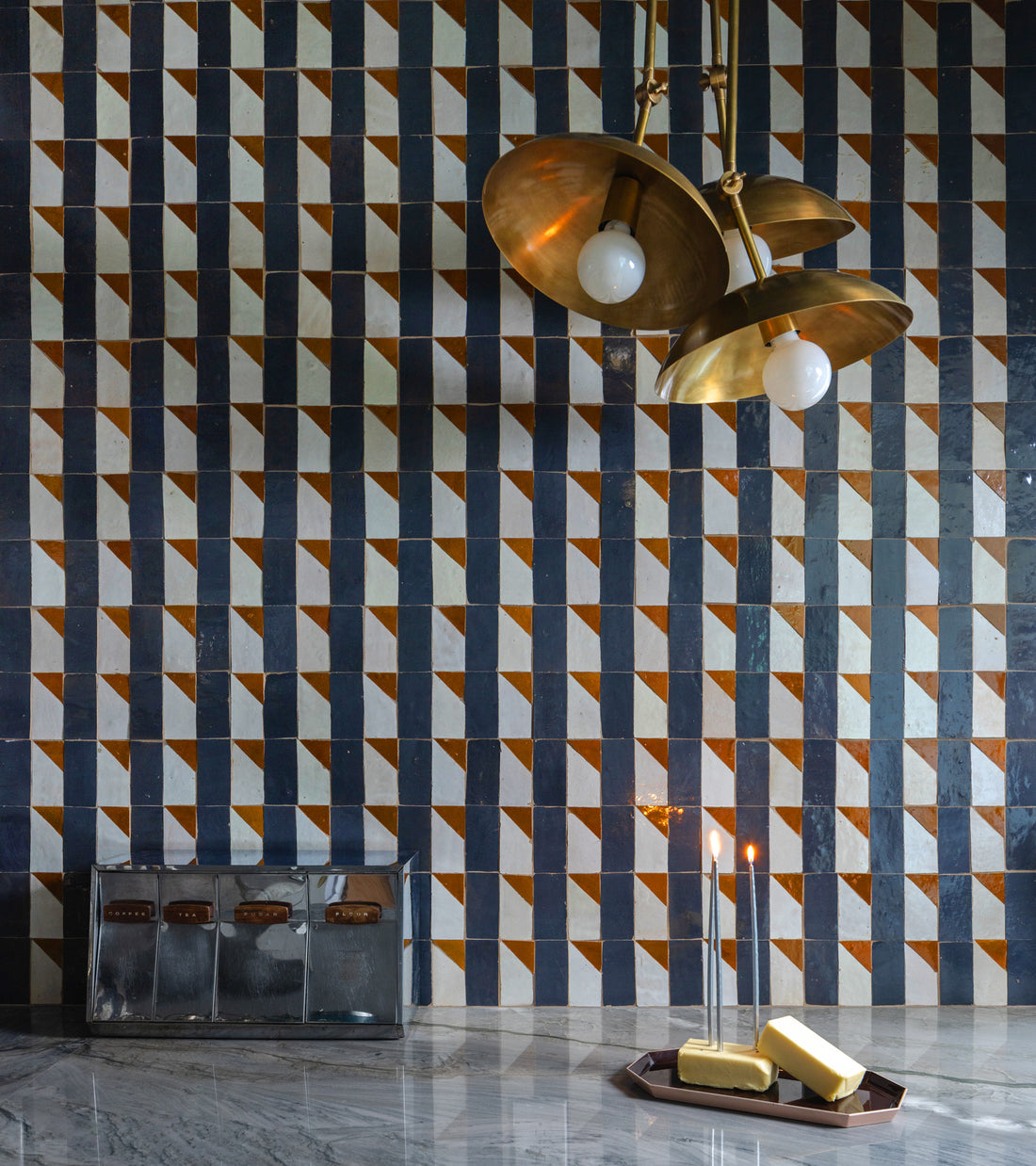 A Radian Inline 2 geometric kitchen backsplash by Zia Tile sets the scene, paired with a gold three-bulb pendant, marble countertop, two lit candles, a tray with gold bars, and a vintage metal box with leather straps in the foreground.