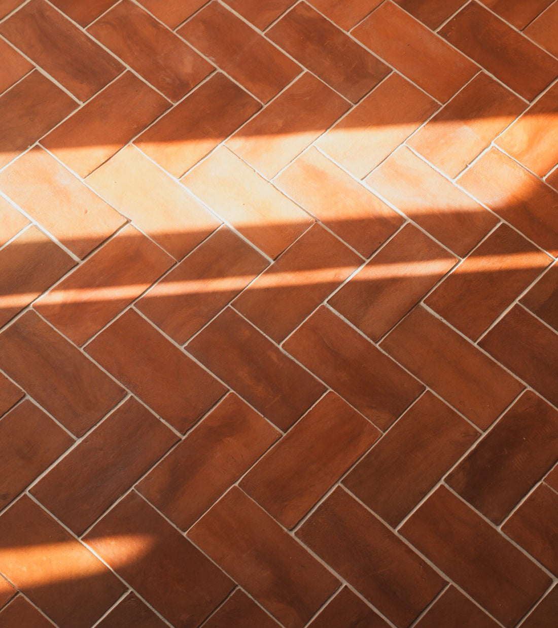 Close-up of Zia Tile’s 6.5x13 Rectangle + Red Clay tiles in a herringbone pattern, sunlight casting diagonal stripes of light and shadow over the rich reddish-brown surface.