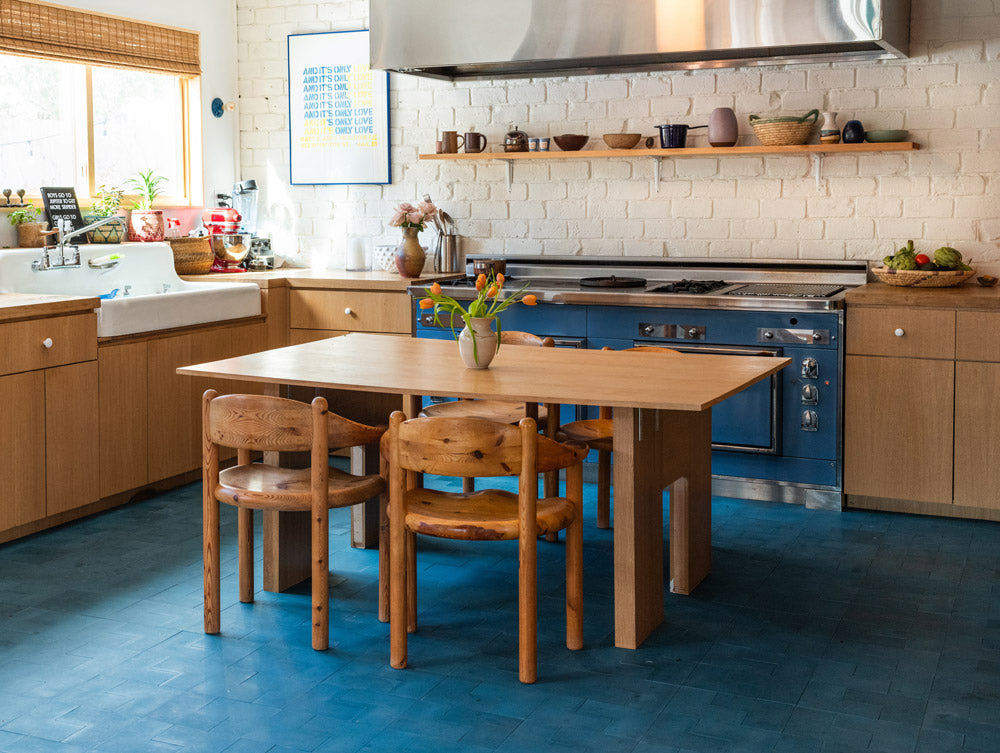 A cozy kitchen with wooden cabinets, a blue stove, and blue tile floor. A wooden dining table with four matching chairs sits in the center, with a vase of flowers on top. Natural light streams in from a window.