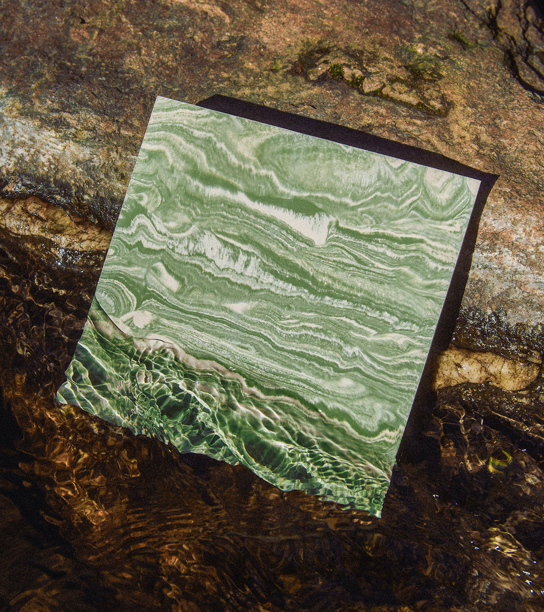 The River Olivine + Bone 8x8 tile by Zia Tile, with its green marble-like surface, rests on a rock near clear brownish water as sunlight casts rippling reflections across the tile.
