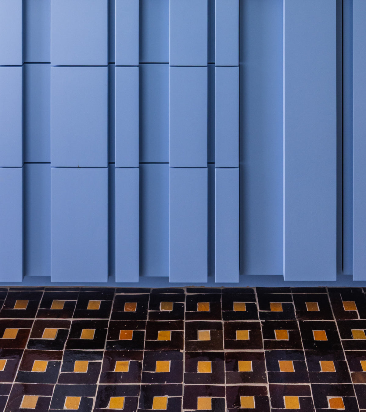 Rubric 5 wall panels by Zia Tile in blue rectangles of varying widths are arranged vertically above a dark, Moroccan-inspired geometric floor with black squares and yellow rectangles.