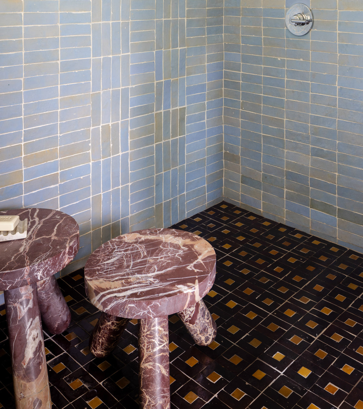 Two round marble stools and a rectangular marble table rest on a dark geometric tiled floor. The walls feature Zia Tile’s Rubric 5, pale blue Moroccan tiles arranged in rectangles with visible grout lines.