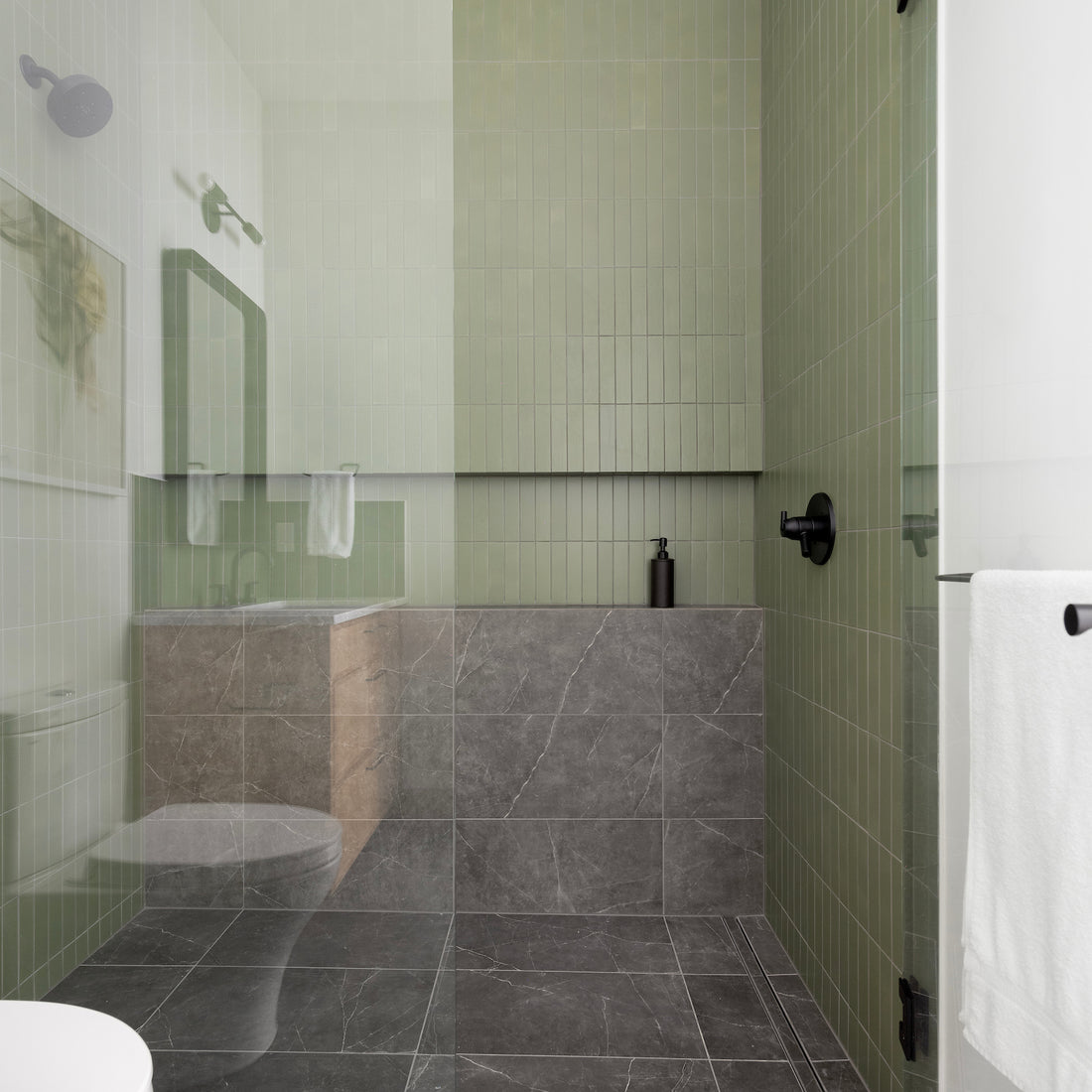 Modern bathroom with green tiled walls, a walk-in glass shower, black fixtures, and dark gray marble floor tiles. A wood vanity with a mirror and towel, and a white towel hangs on the wall. Minimalist and clean design.