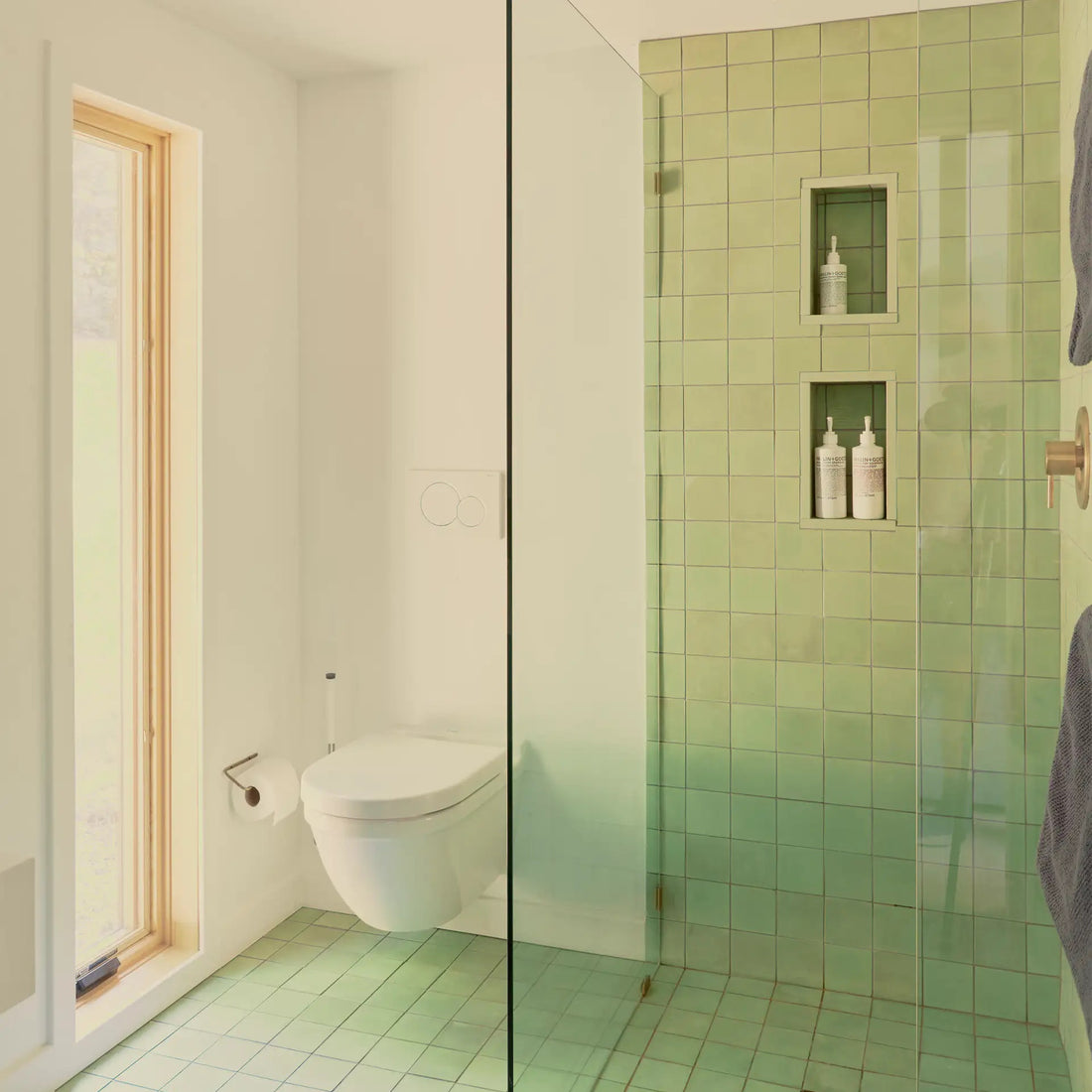 Modern bathroom with green tile floor and shower walls, glass shower divider, built-in shower shelves holding bottles, wall-mounted toilet, wooden framed window, and natural light filling the space.