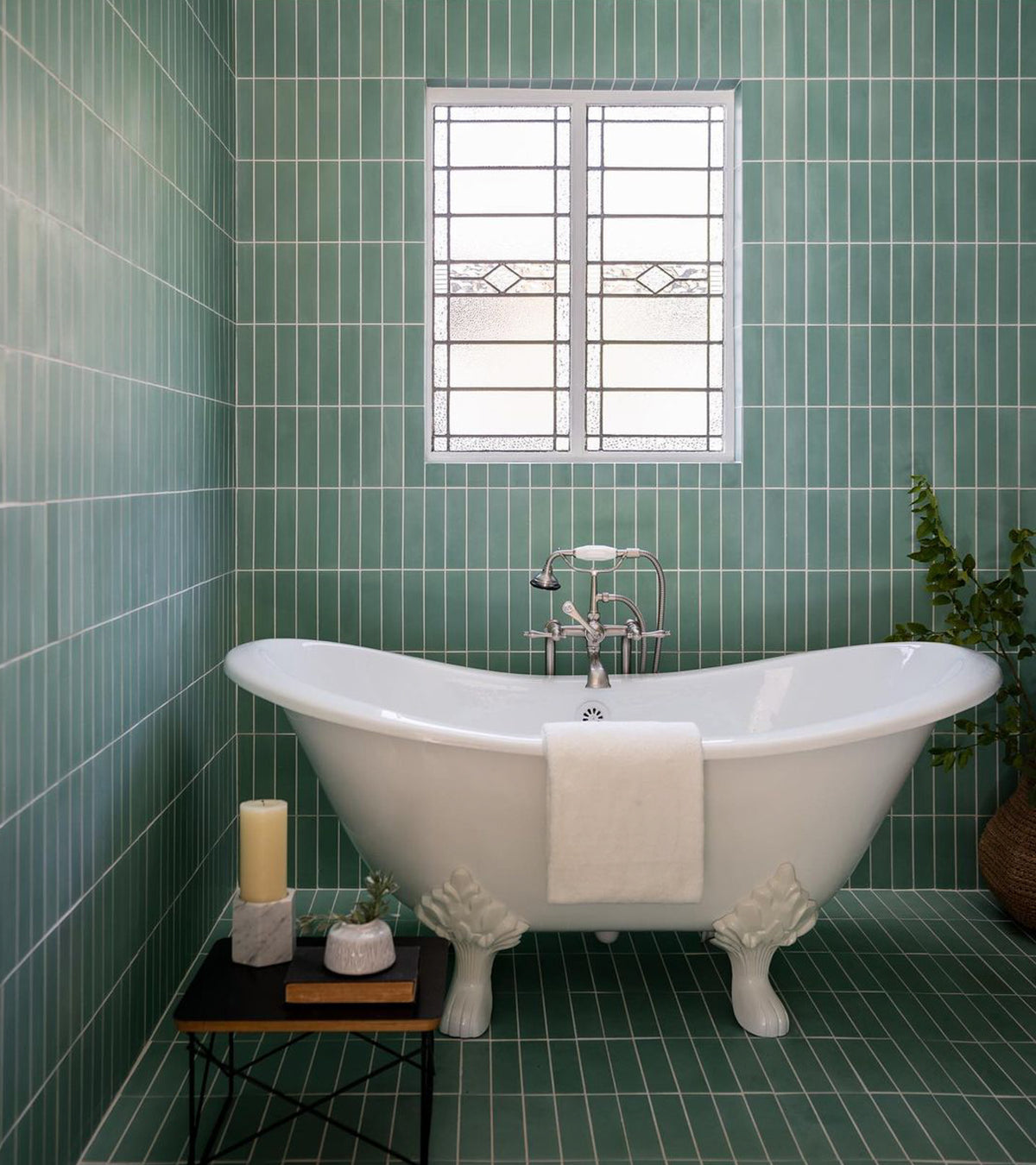 A white clawfoot tub with a towel rests in a bathroom featuring vendor-unknown's Salvia 2x8 green subway tile. A small table with candles sits nearby, and a frosted window above the tub lets in light.