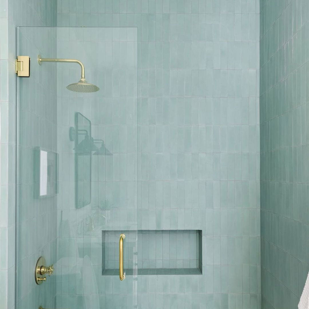 A modern shower with pale green rectangular tiles, a built-in wall niche, a glass door with a brass handle, and a brass showerhead and faucet.