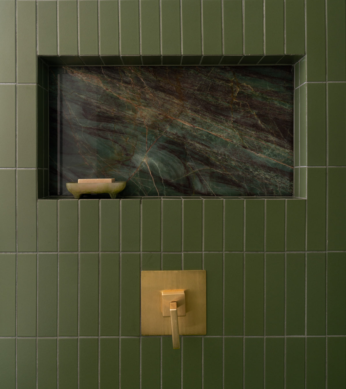 A shower wall showcases Sequoia 2x8 sage green matte ceramic tile by Zia Tile, featuring a niche lined with dark green marble. Inside is a small soap dish, and below sits a gold shower handle on a square backplate.