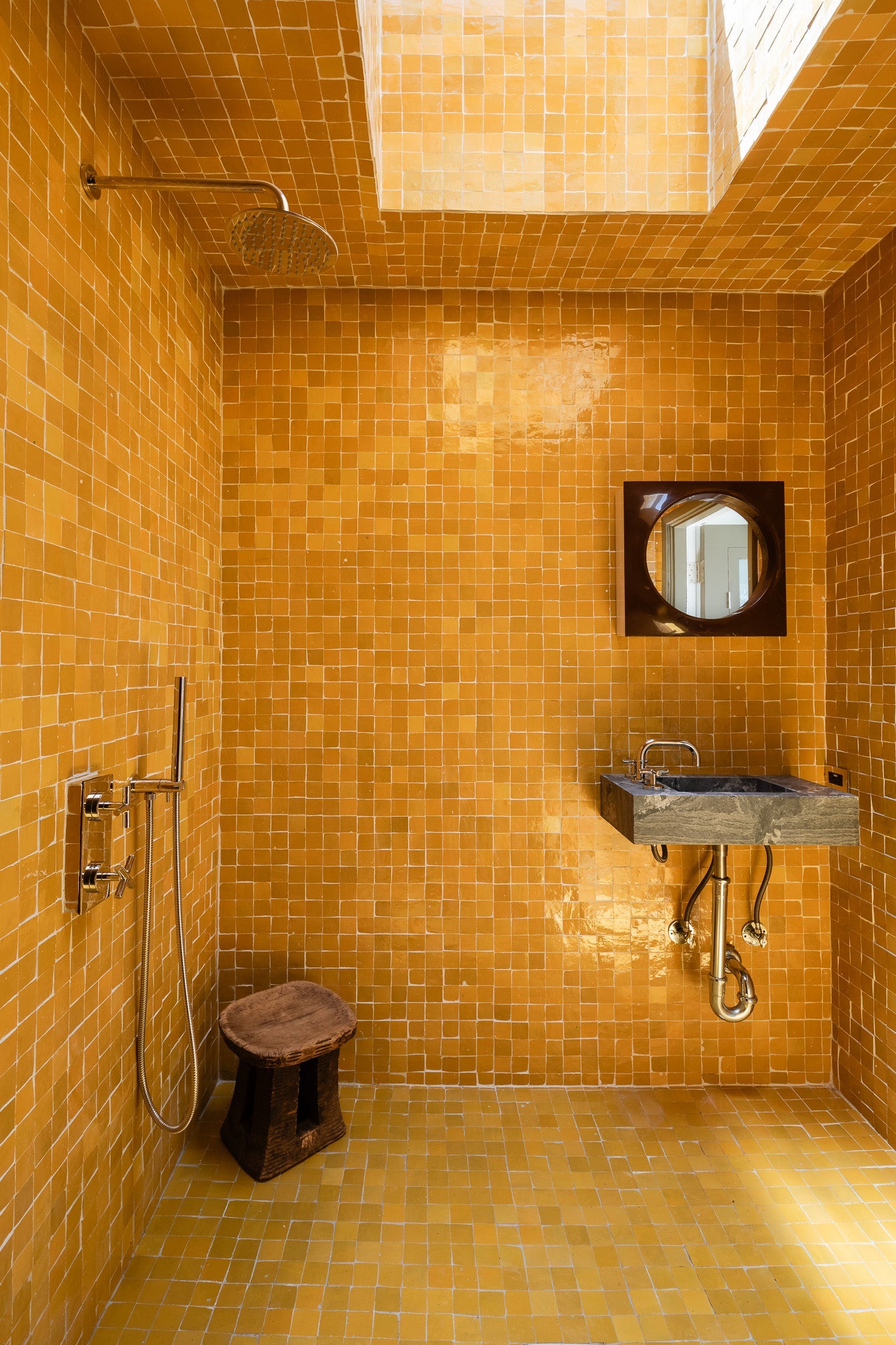 A bathroom with glossy orange tiles, a rainfall shower, a small wooden stool, a gray sink with exposed pipes, and a round mirror on the wall, lit by natural light from a skylight above.