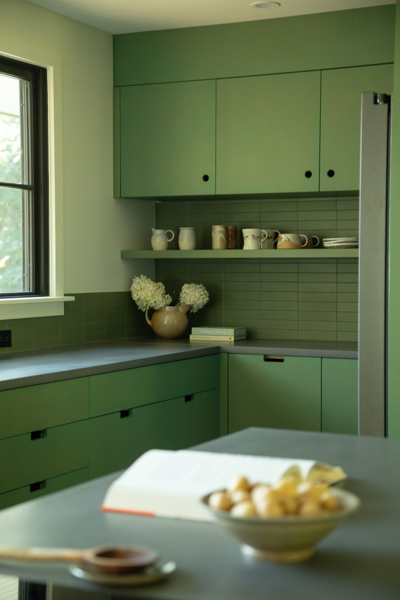 A modern kitchen with matte green cabinets, gray countertops, and open shelves displaying mugs, pitchers, and a vase with white flowers. A book and bowl of potatoes sit on the counter in the foreground.