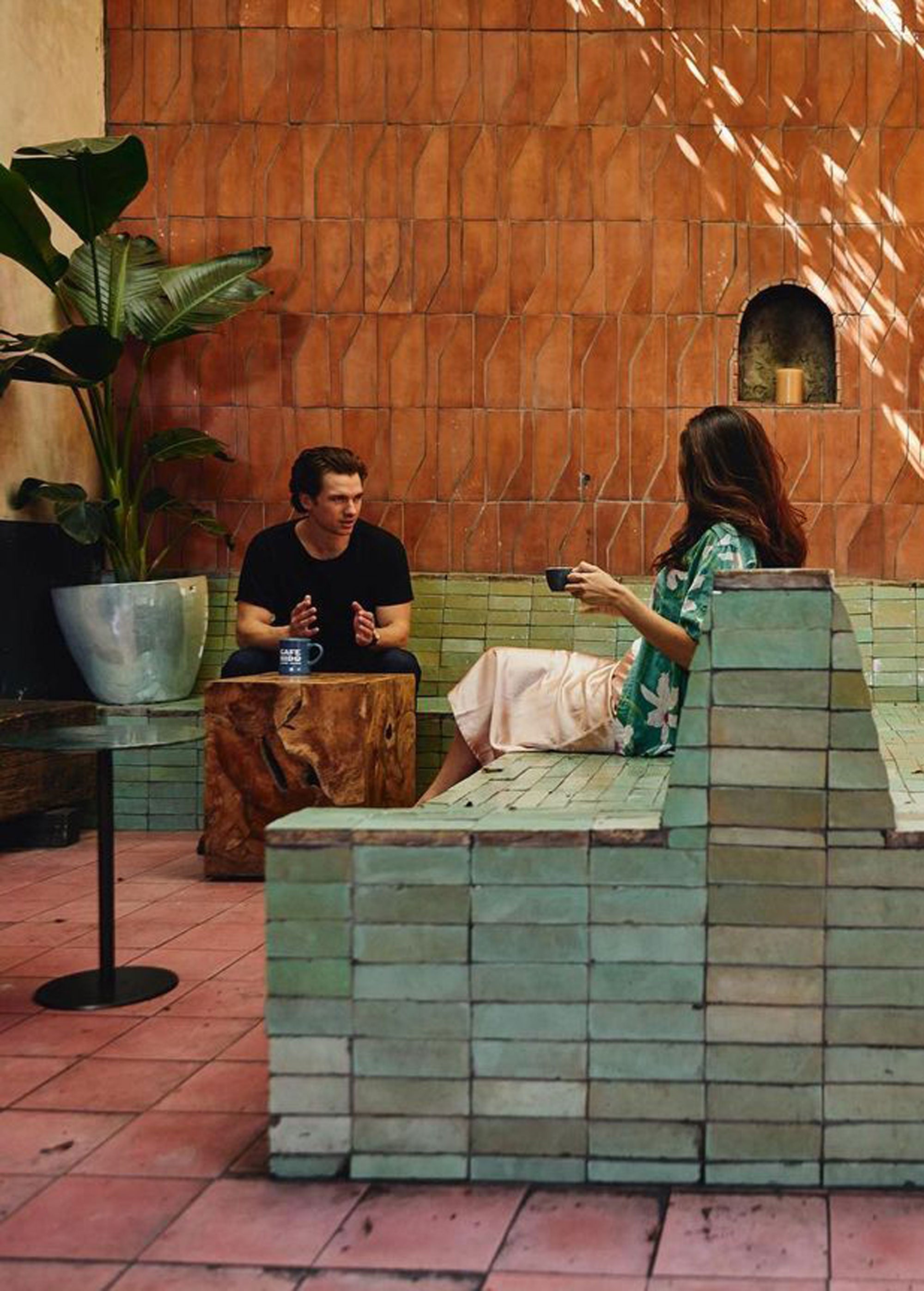 A man and a woman sit across from each other in a stylish, tiled outdoor space with pink floors and green benches. The woman holds a cup, and there are potted plants and sunlight streaming over the terracotta wall.