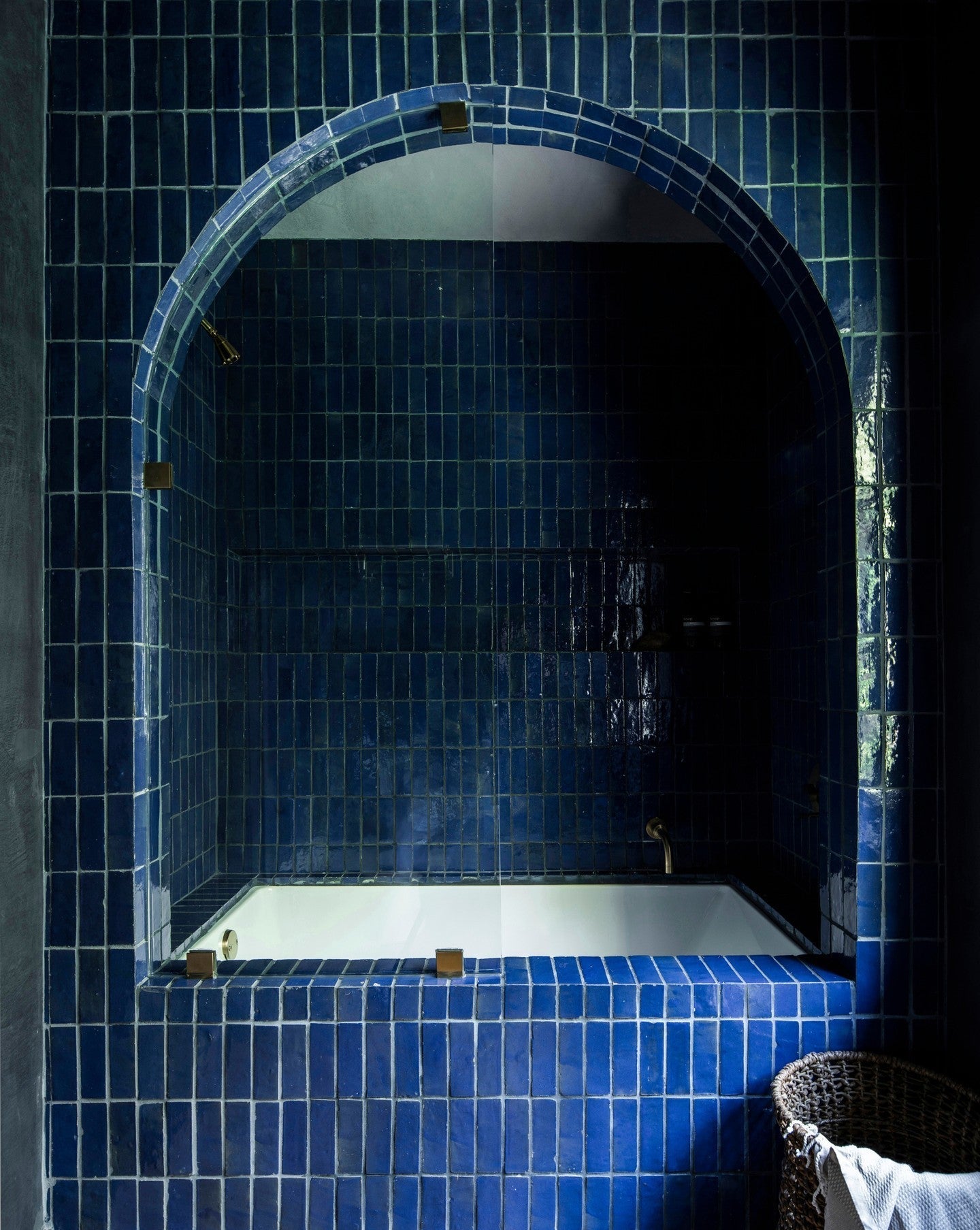A bathtub is set within an arched alcove, both covered in glossy dark blue tiles. A woven basket sits next to the tub, and the overall lighting is dim, creating a moody atmosphere.