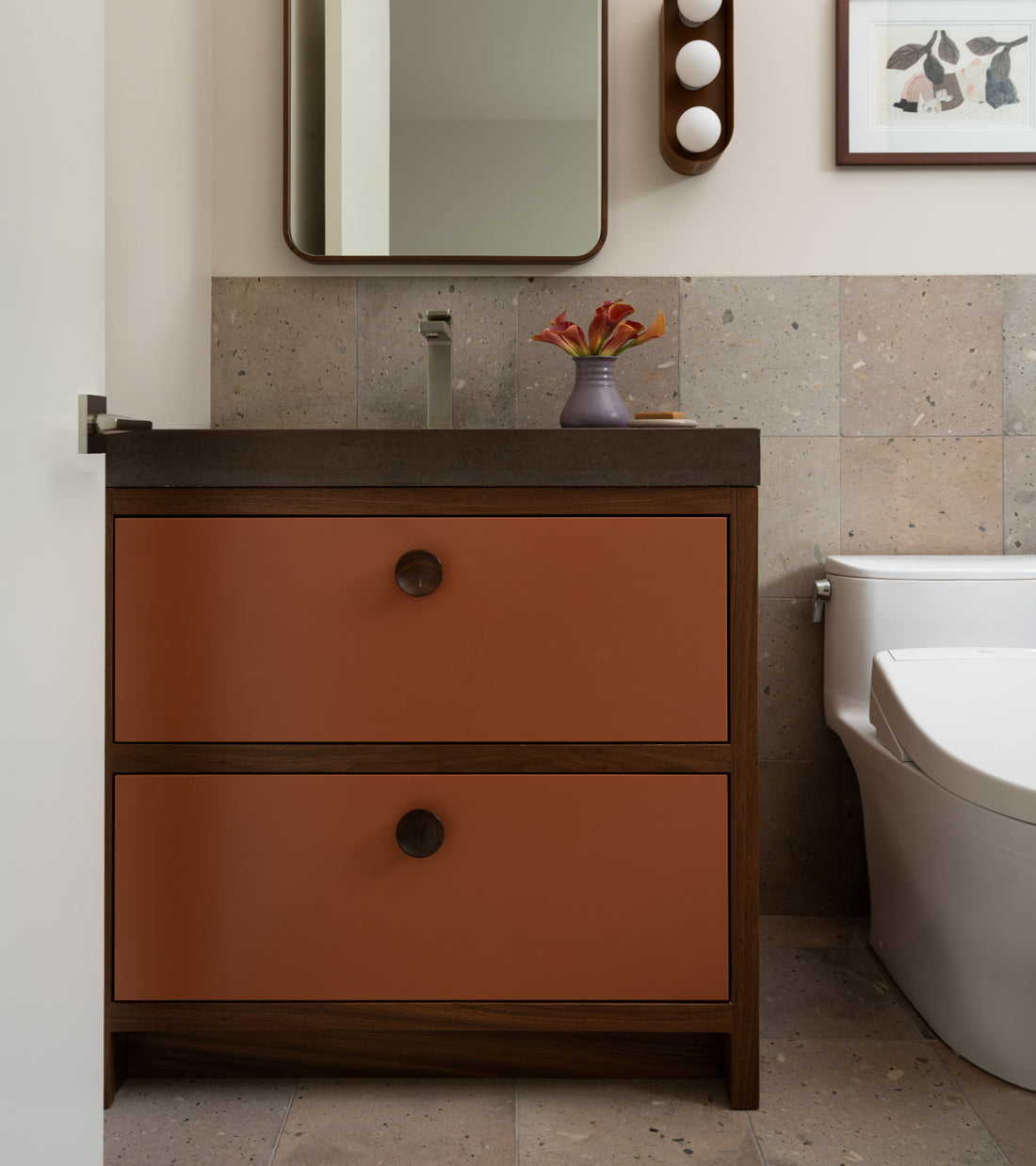 A modern bathroom features a vanity with two rust-colored drawers, a dark countertop, a vase of flowers, a rectangular mirror, and a white toilet against beige Sierra 12x12 tile walls by Zia Tile.