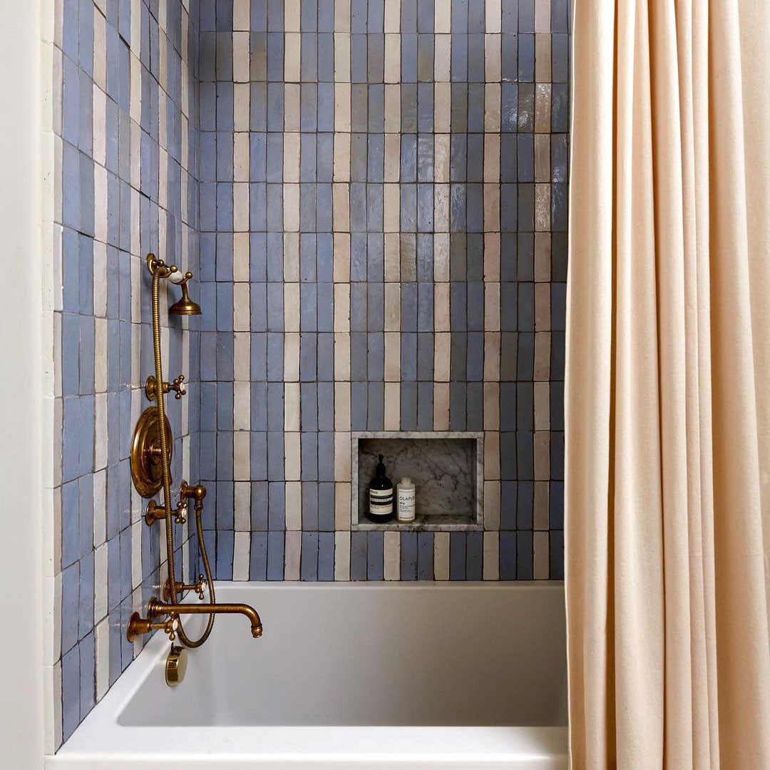 A bathtub with gold fixtures is set against a wall of vertical blue and white tiles. A recessed shelf with toiletries sits in the tiled wall, and a beige shower curtain is pulled partway closed.