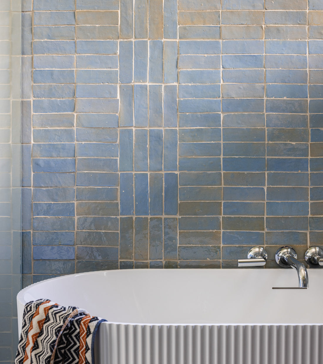 A modern bathroom features a white tub with a chrome faucet and a zigzag-patterned towel, set against a blue, rectangular-tiled wall using Zia Tile's Skylight 2x6 tiles.