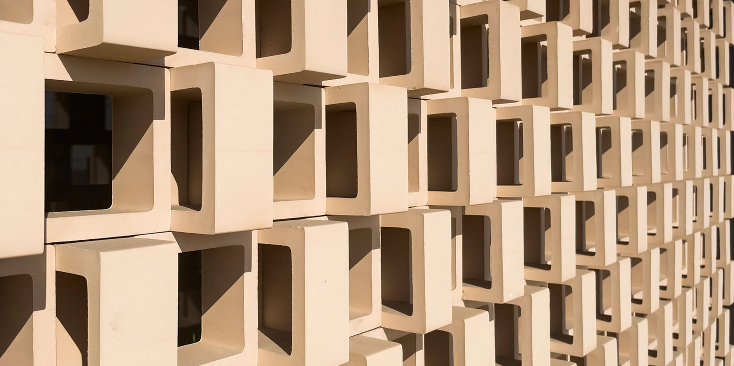 A close-up view of a beige, geometric architectural wall made of repeating rectangular openings, creating a pattern of light and shadow.