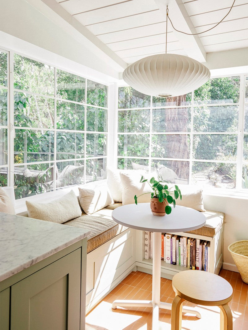 A bright, sunlit breakfast nook with large windows, a built-in bench with cushions, a round white table with a potted plant, a wooden stool, and books stored beneath the bench. Lush greenery is visible outside.