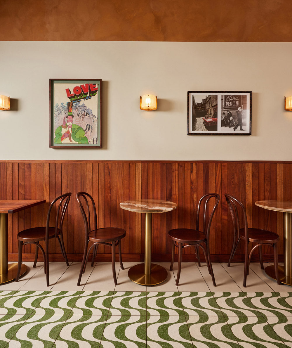 A cozy cafe interior with wooden chairs, round tables, wood-paneled walls, framed art and photos, wall sconces, and green-and-white wavy patterned floor tiles.