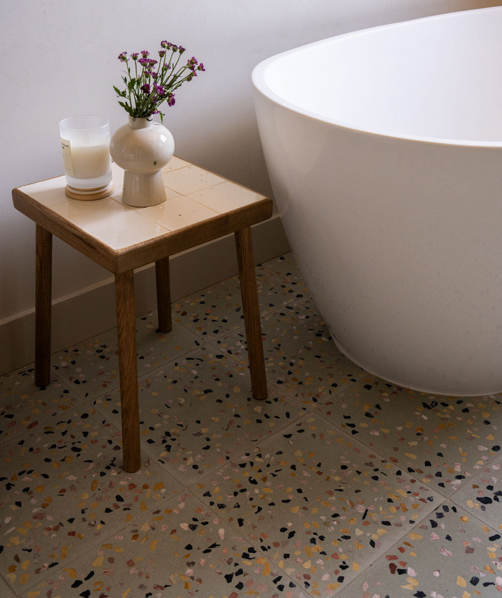 A small wooden table with a candle and a vase of flowers stands next to a white bathtub on a terrazzo tile floor in a modern bathroom.