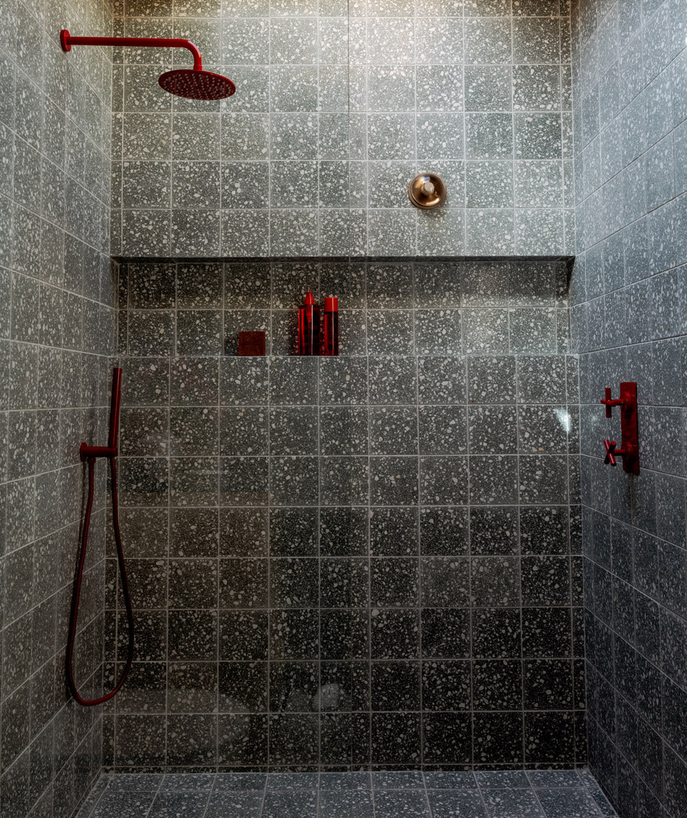 Modern shower with dark speckled tiles, a red rain showerhead, red handheld shower, red controls, and a recessed shelf holding red bottles and a sponge. Light streams in from above.